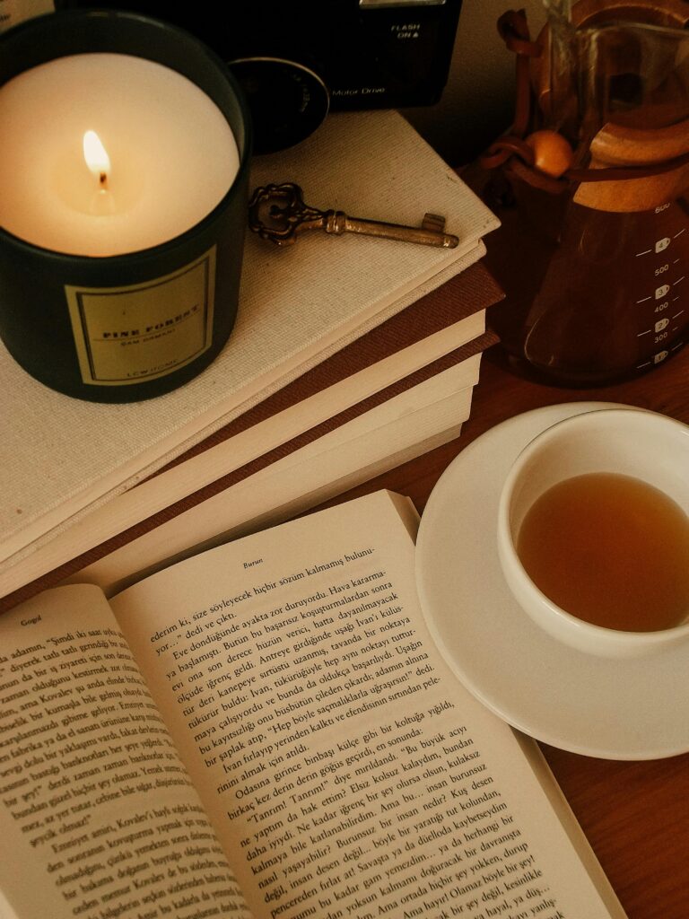 A cozy scene with an open book, a lit candle, a vintage key, stacked books, and a cup of tea on a wooden table.